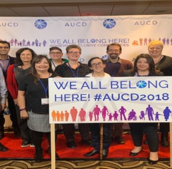 11 IDHD staff, faculty, and alumni pose in front of a sign which reads 