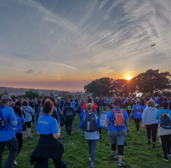 A large group of people people wearing blue T-shirts walking on the grass towards sunset over the ocean behind 3 trees.
                  