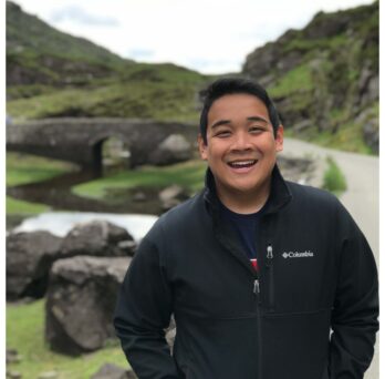 Alan Sadural is wearing a black jacket and standing outdoors with a very verdent landscape in the background
                  