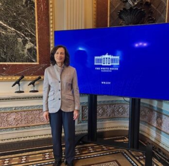 (ID: woman with dark hair wearing gray/black business suit standing next to large monitor showing white house logo.  Behind is shown marbled walls.
                  