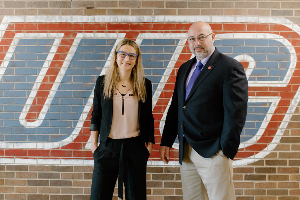 Karrie Hamstra-Wright and John Coumbe-Lilley standing in front of brick wall with UIC logo