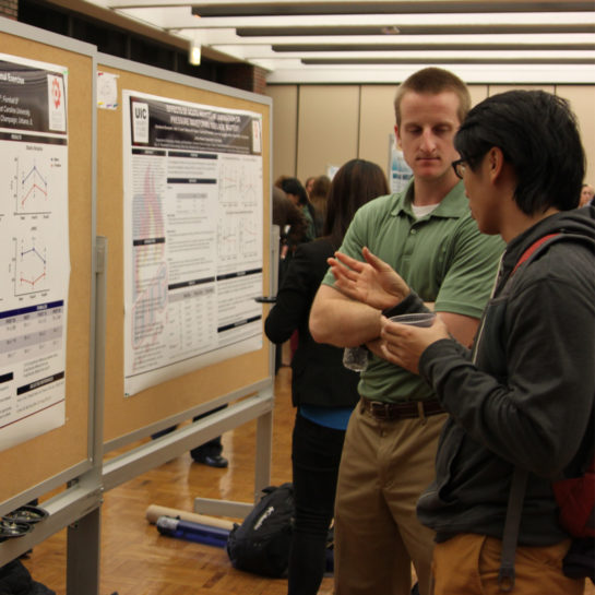 Two men talk as they stand before a research poster