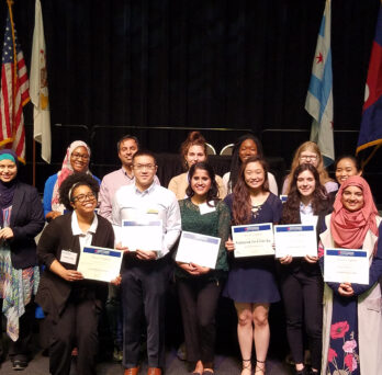 A group of fifteen student winners stand in two rows holding their award certificate
                  