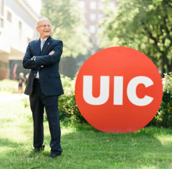 Carlos Crespo standing in the west side of the UIC campus next to a large red UIC circle mark
                  