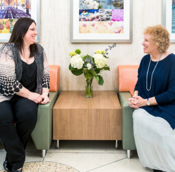 AnnaMaria Baraglia and Carol Gottlieb smile at each other while sitting next to one another
                  