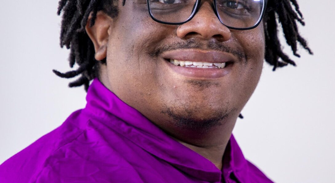 A professional head shot of Timotheus “T.J.” Gordon Jr wearing dark-rimmed glasses and a purple long-sleeved shirt