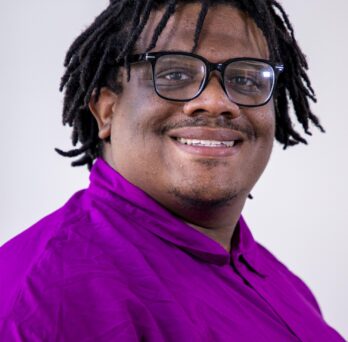 A professional head shot of Timotheus “T.J.” Gordon Jr wearing dark-rimmed glasses and a purple long-sleeved shirt
                  