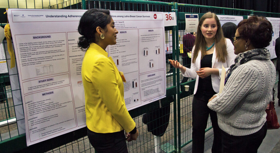 Two undergraduate students stand in front of their research poster while one of them explains the posters findings to a woman who is facing them.