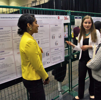 Two undergraduate students stand in front of their research poster while one of them explains the posters findings to a woman who is facing them.
                  