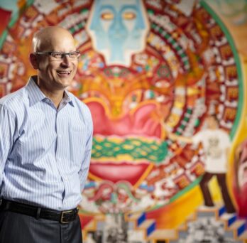 Carlos Crespo stands in front of a colorful wall mural
                  