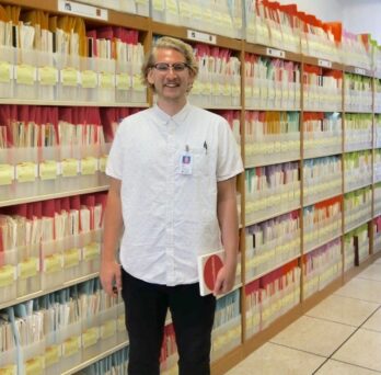 PhD student Drew standing in front of bookshelves of documents
                  