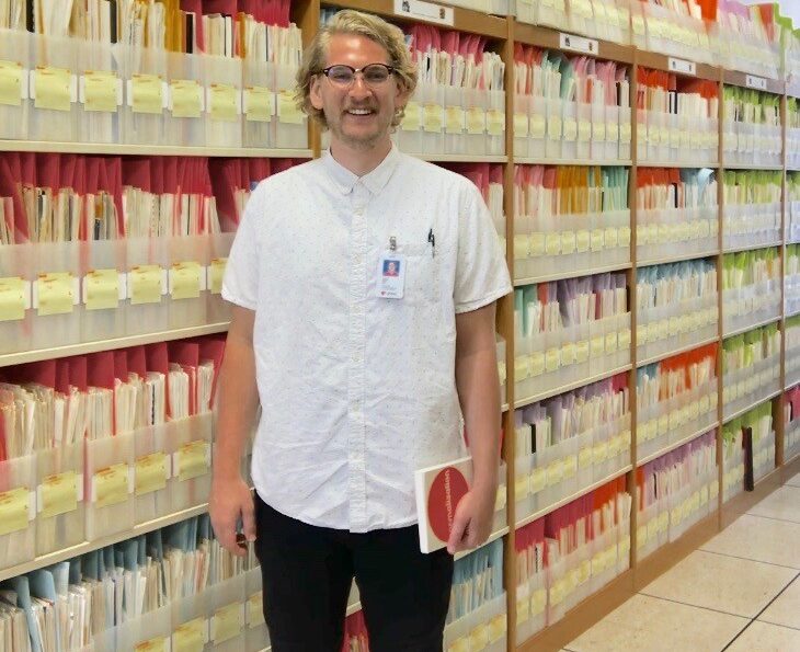 PhD student Drew standing in front of bookshelves of documents