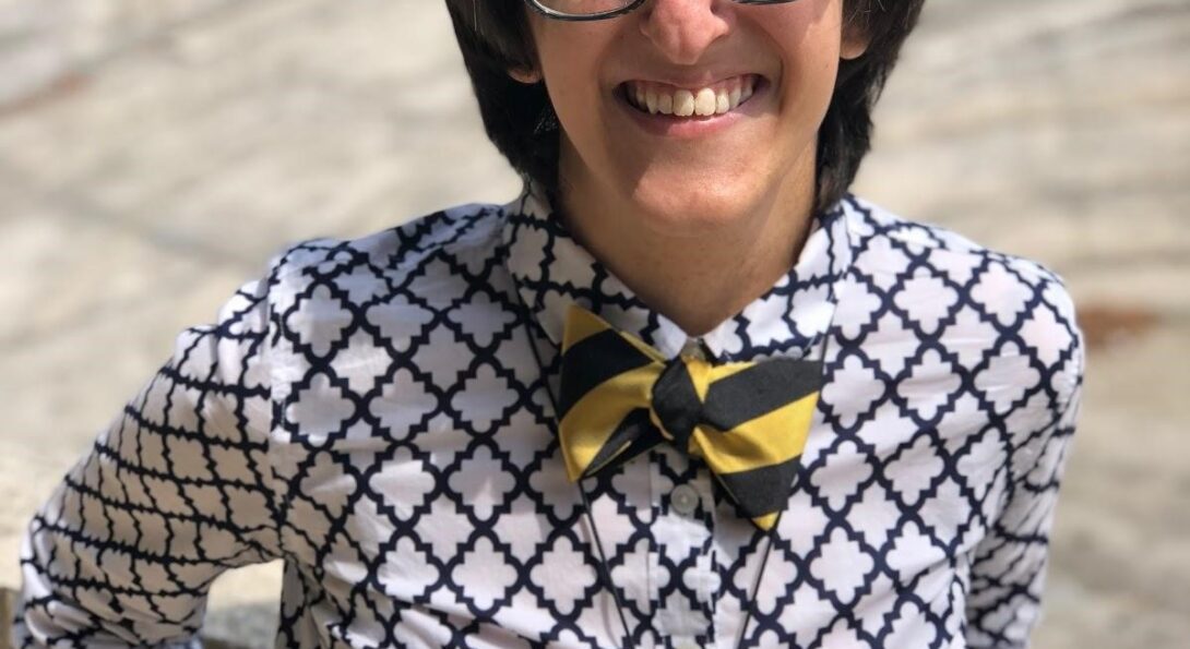 A female located outside. The short-haired brunette woman is wearing a white and blue patterned button-down shirt and a black-yellow striped bow tie. She is also wearing glasses and is smiling widely while squinting her eyes