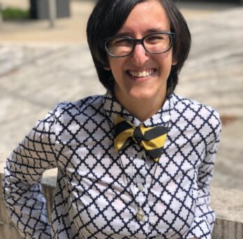 A female located outside. The short-haired brunette woman is wearing a white and blue patterned button-down shirt and a black-yellow striped bow tie. She is also wearing glasses and is smiling widely while squinting her eyes
                  