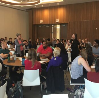 Tina Chase leading the post-role playing exercise discussion in a room full of students sitting around round tables
                  