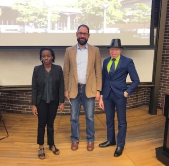 Esther Allen Nakamwa, Rob Gould and Peter Ogik standing together in front of a classroom whiteboard/projection screen
                  