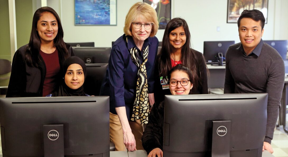 Valerie Prater standing in the middle of a group of two students who sit behind desktop computers and three students who are standing beside her