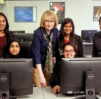 Valerie Prater standing in the middle of a group of two students who sit behind desktop computers and three students who are standing beside her
                  