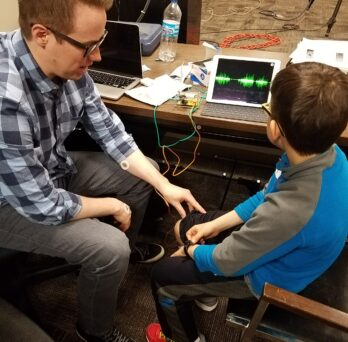 An AHS researcher and elementary school-aged student sit in front of health monitoring equipment
                  