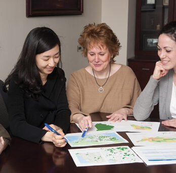 Jenna Heffron stitting with a group at a table looking at papers
                  