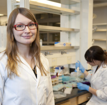 Krista Varady wears a white lab coat while standing in a research laboratory
                  