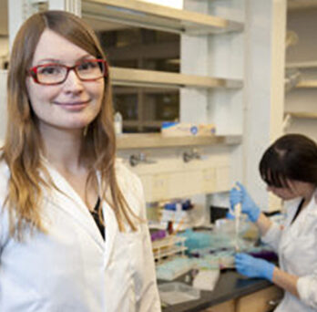 Krista Varady in a white lab coat faces the camera. A person wearing a white lab coat is in the background.
                  