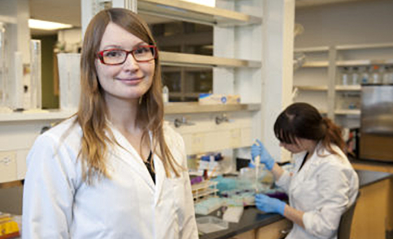 Krista Varady in a white lab coat faces the camera. A person wearing a white lab coat is in the background.