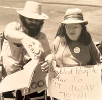 Larry Voss and Carol Gill at a protest, both sitting in wheelchairs. Carol Gill is holding a sign that says 