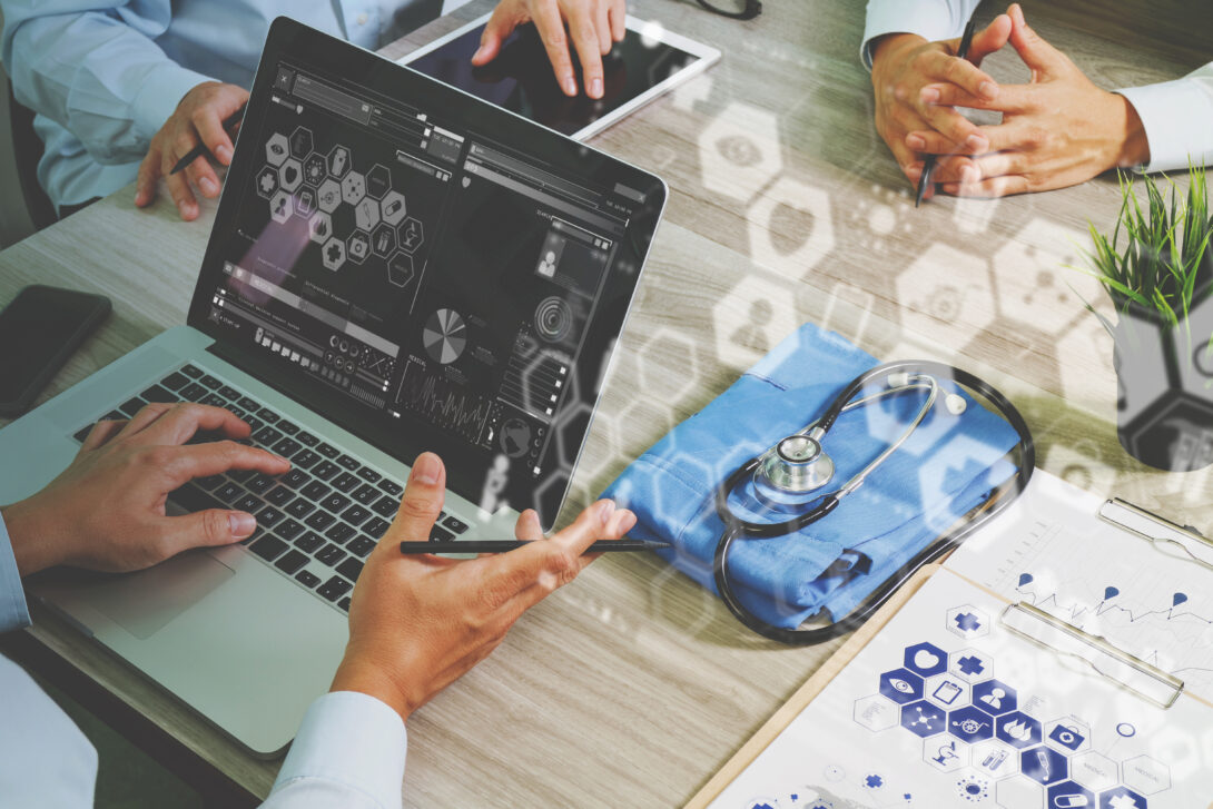 A laptop is being used by an individual who is sitting at a square table with two other individuals, one of which is using a tablet. A stethoscope is on the table. Transparent health related graphics are superimposed on the image.