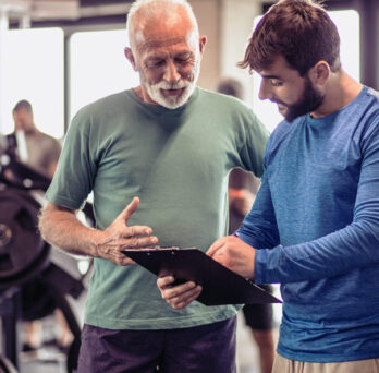 Two men in a clinical gym setting examine a tablet together, discussing fitness routines or workout plans.
                  