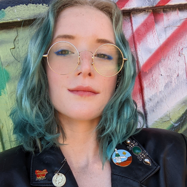 White women wearing gold round glasses with blue/green hair.