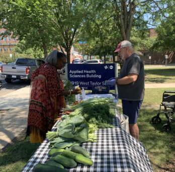 Jim (right), a volunteer from Just Roots Chicago, helps Theresa (left), a UIC OT Faculty Practice patient, stand outisde in front of 1919 W. Taylor St. where a long table is set up with fresh produce. 