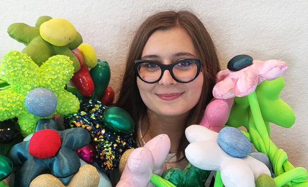 A white woman with thick cat eye glasses smiles into the camera. She sits in front of three large sequin smiley faces and is wearing a shirt with more smiley faces on it.