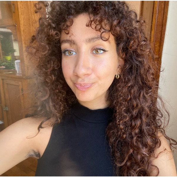 Nour holds up her phone to take a picture of herself. She is wearing a black tank top with her brown curly hair down, and she smiles into the camera.