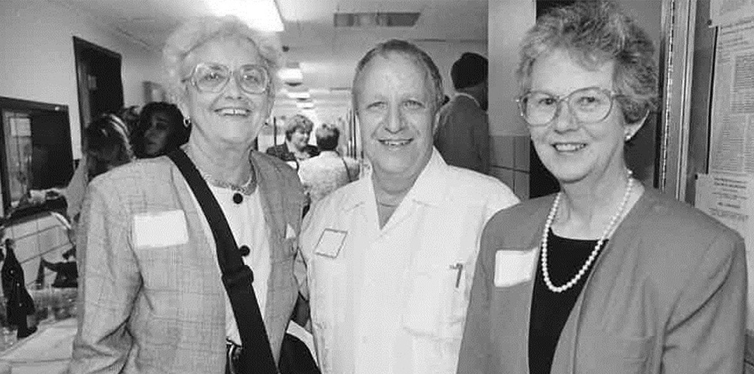 A group of 3 people, two women and one man, standing and smiling at the camera with name tags on. Black and white photo.