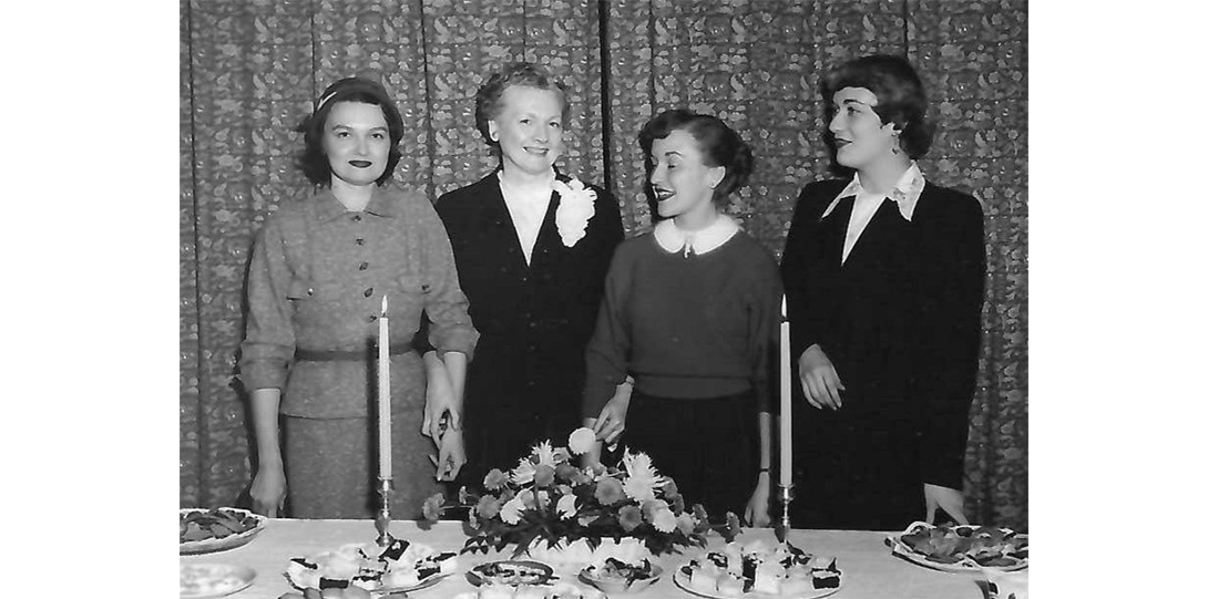 A group of four women in a black and white photo standing in front of a table with flowers.