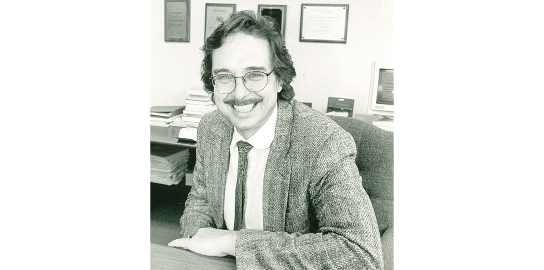 A black and white photo of a man sitting in an office behind a desk, smiling at the camera