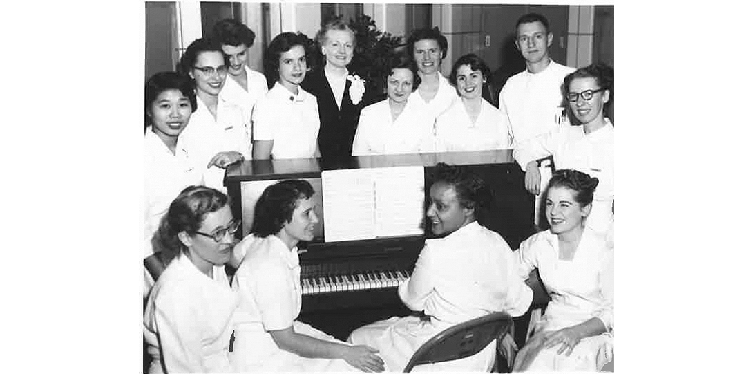 A black and white photo of a group of women gathered around a piano