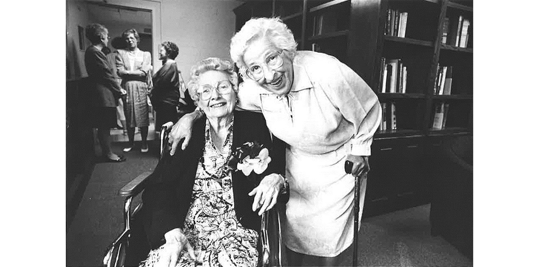 Two women smiling at the camera, one is seated in a wheelchair and one uses a cane.