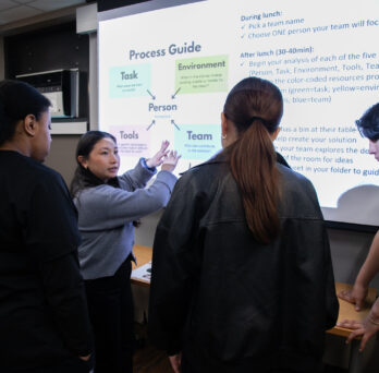 A group of students gather around a projection screen in a classroom
                  