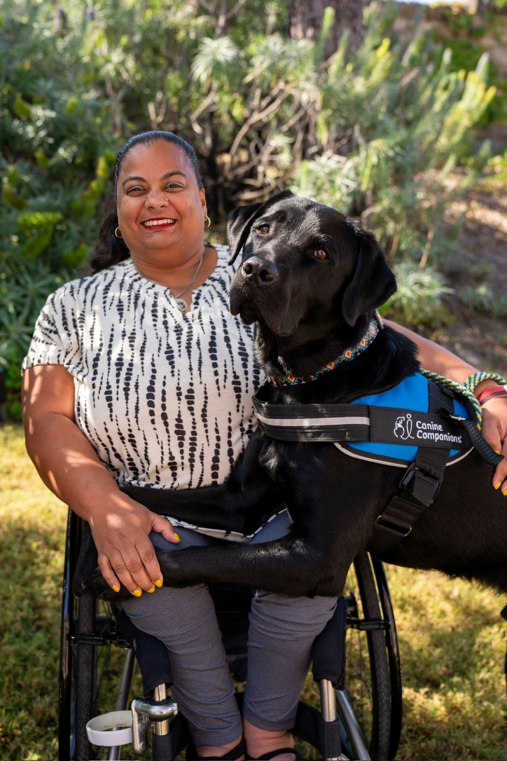A woman sits in a wheelchair with a black service dog beside her.